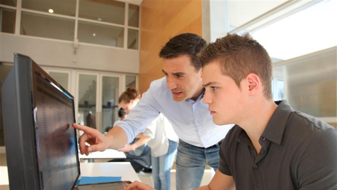 jaunietis sēž pie datora klasē, blakus stāvošais vīrietis rāda kaut ko viņam monitorā 