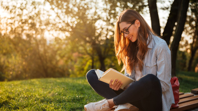 studente ar brillēm sēž parkā uz soliņa un lasa grāmatu