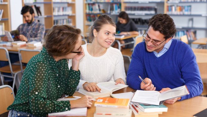 Erasmus+ TCA; trīs studenti sēž bibliotēkā pie viena galda strādā