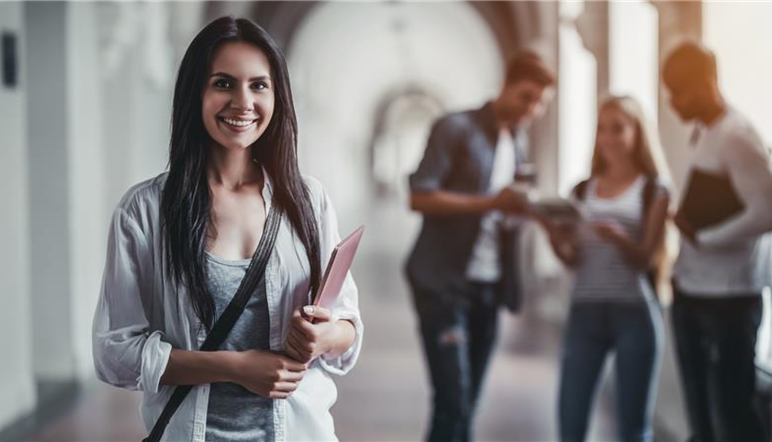 smaidīga studente stāv priekšplānā, rokās tur grāmatas, aizmugurējā plānā stāv citi studenti
