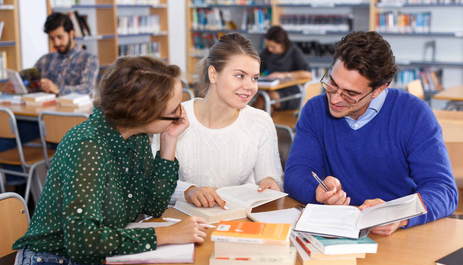Erasmus+ TCA; trīs studenti sēž bibliotēkā pie viena galda strādā
