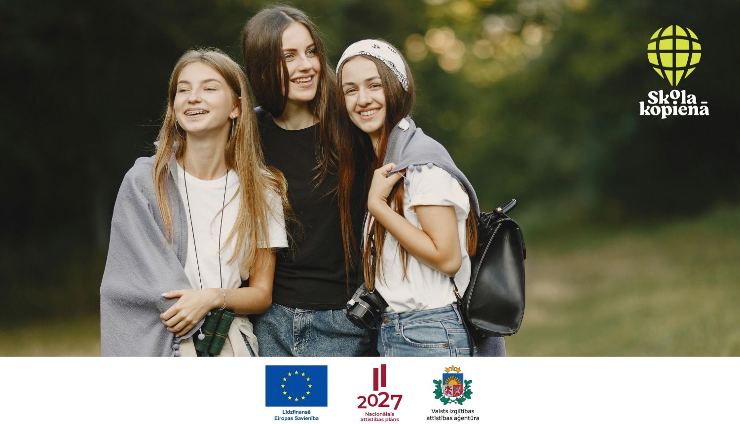 attēlā trīs jaunas meitenes stāv kopā un smaida, fonā zaļi koki, zāle; stūrī Skola-kopienā logo