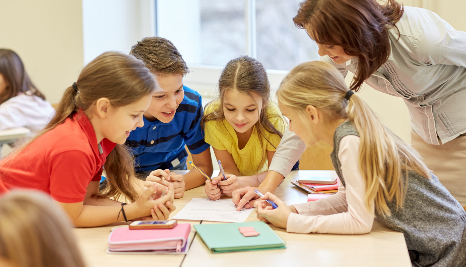 skolotāja palīdz skolēniem pildīt uzdevumu, četri skolēni sēž pie galda grupā pilda uzdevumu