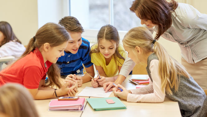 skolotāja palīdz skolēniem pildīt uzdevumu, četri skolēni sēž pie galda grupā pilda uzdevumu