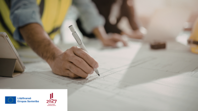 vīrietis atstarojošā darba vestē atzīmē kaut ko ar pildspalvu uz lielā būvdarbu rasējuma, kas stāv uz galda