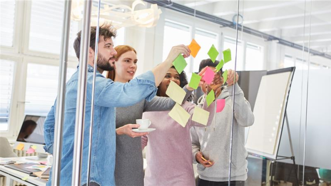 cilvēku grupa stāv pie stikla sienas un līmē krāsainas piezīmju lapiņas