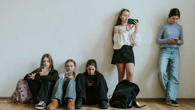 pusaudži sēž uz grīdas un stāv pie sienas visi skatās telefonos