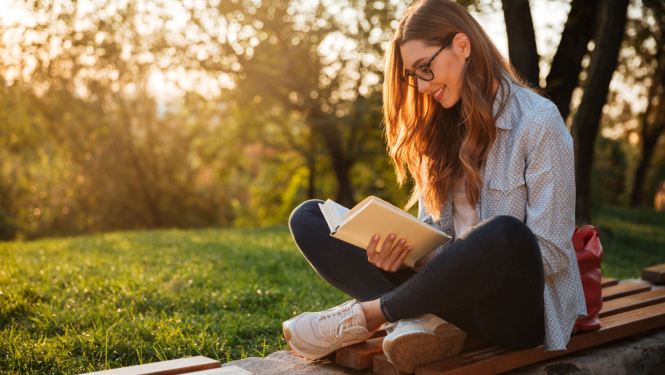 studente ar brillēm sēž parkā uz soliņa un lasa grāmatu