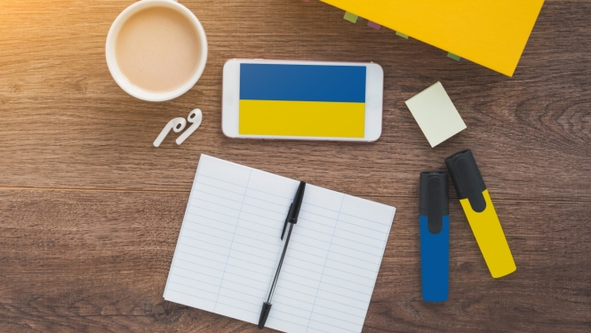 Ukrainas karogs mobilā telefona ekrānā, kas nolikts uz galda, blakus darba klase ar pildspalvu un divi flomasteri dzeltenā un zilā krāsā