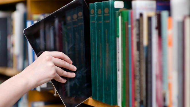 cilvēka roka stiepjas ar planšeti to ievietot grāmatu plauktā bibliotēkā