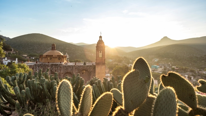 Meksikas dabas skats, priekšplānā kaktusi, aizmugurē kalni un baznīca