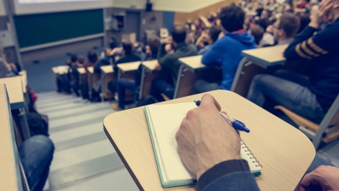 studenti sez lielā augstskolas auditorijā priekšplānā vīrieša roka ar pildspalvu un uz galda klade