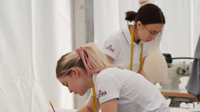 Meitene pie galda zīmē ar zīmuli, fonā redzama otra konkursante ar mērlenti ap kaklu