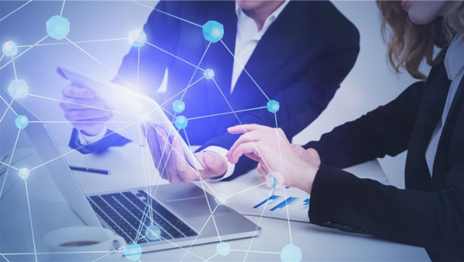 sieviete un vīrietis uzvalkā sēž pie darba galda, uz galda atvērts portatīvais dators, kopā skatās vienā izdrukātā dokumentā