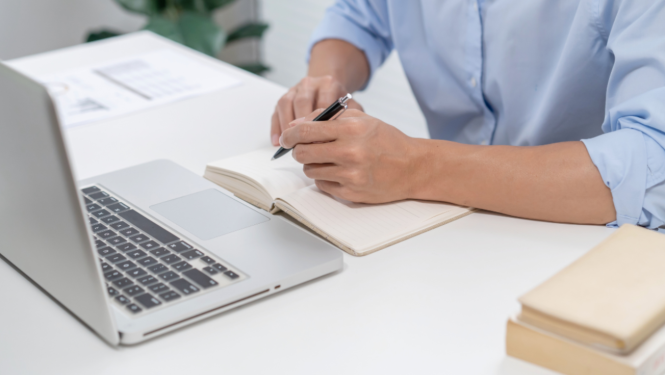 Cilvēks sēž pie galda ar portatīvo datoru un piezīmju bloknotu