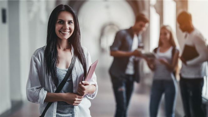 smaidīga studente stāv priekšplānā, rokās tur grāmatas, aizmugurējā plānā stāv citi studenti