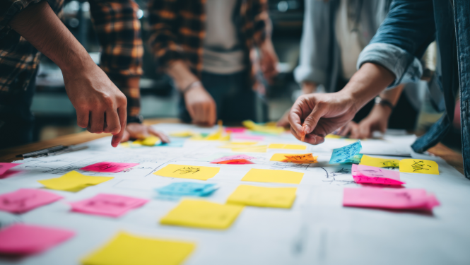 vairāki cilvēki pie galda veido kopīgu projektu, lipina krāsainas lapiņas uz liela papīra