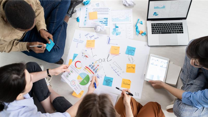 vairāki cilvēki šēž pie darba galda pa apli, uz galda ir klēpjdators un dažādas pierakstu lapas