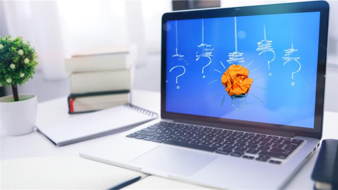 portatibvais dators stāv uz galda, blakus grāmatu kaudzīte, uz ekrāna ir stilizēta lampiņa