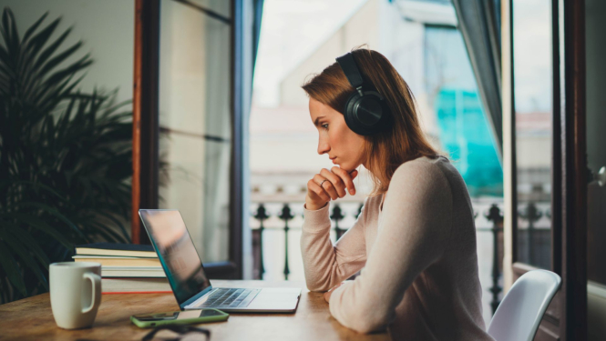 jauna sieviete sež pie galda ar austiņām uz galvas, priekšā viņai uz galda atveērts portatīvais dators