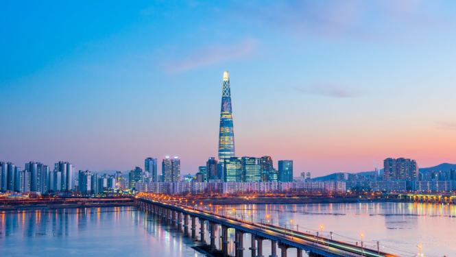attēlā skats uz Korejas galvaspilsētu, priekšā tilts pāri upei, aizmugurējā plānā augšceltne, saulriets