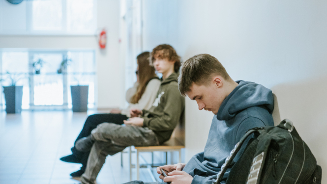 skolēni ar telefonu rokās sēž uz krēsla skolas gaitenī 