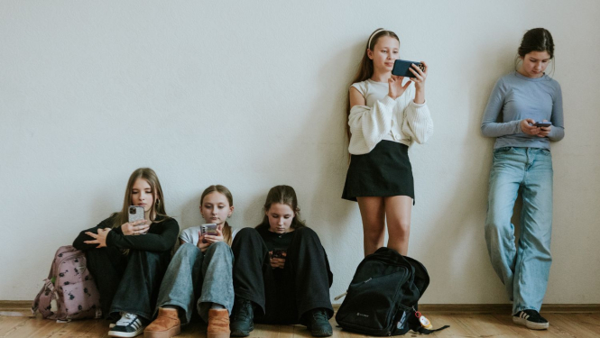 pusaudži sēž uz grīdas un stāv pie sienas visi skatās telefonos