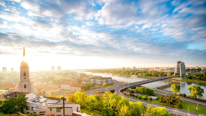 Attēlā skats uz Slovākijas galvaspilsētu, pa kreisi baznīcas tornis, pa visu upe, fonā spoža saule