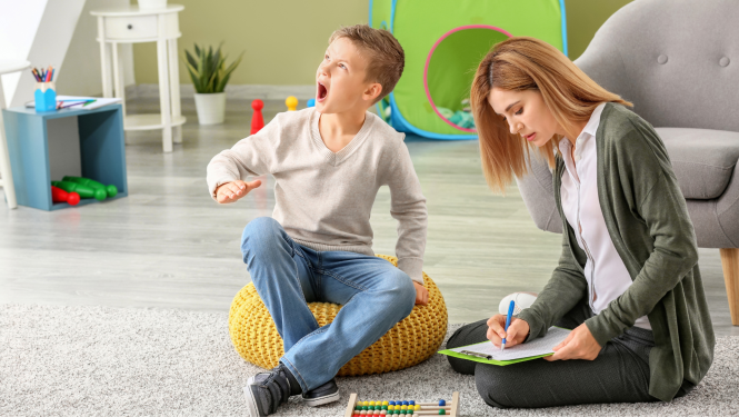 bērns ap 8 gadi sēž uz pufa un bļauj, blakus sēž psiholoģe un pieraksta kaut ko mapītē