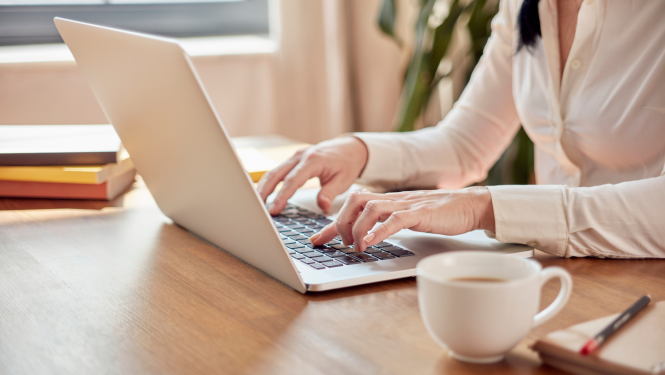 attēlā sieviete sēž pie portatīvā datora raksta uz klaviaraturas, uz galda stāv krūze