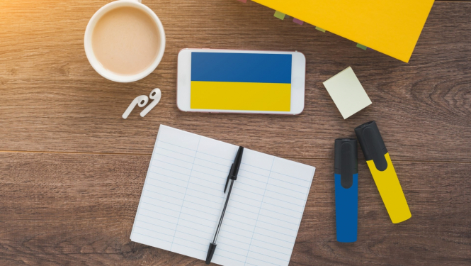 Ukrainas karogs mobilā telefona ekrānā, kas nolikts uz galda, blakus darba klase ar pildspalvu un divi flomasteri dzeltenā un zilā krāsā