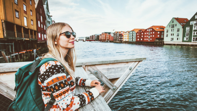 jauna meitene ar mugursomu Norvēģijas pilsētā stāv pie upes 
