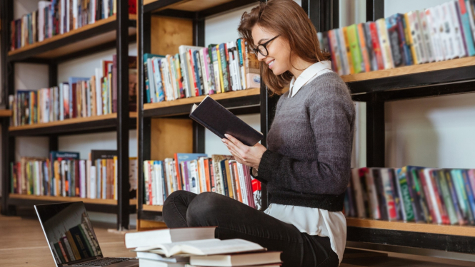 jauna sieviete ar brillēm sēž uz grīdas ar grāmatu rokās biliotēkā
