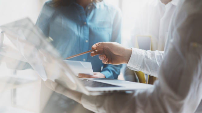 vīrietis baltā kreklā rāda ar zīmuli uz dokumentiem blakus stāv sieviete kreklā