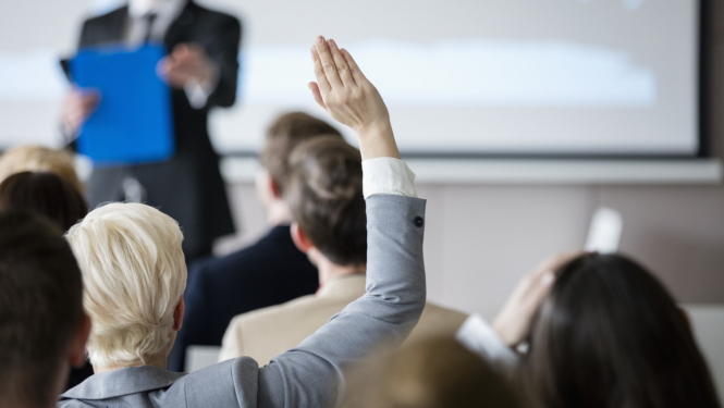 sieviete ceļ augšā roku semināru zālē priekšā stāv vīrietis uzvalkā pie tāfeles