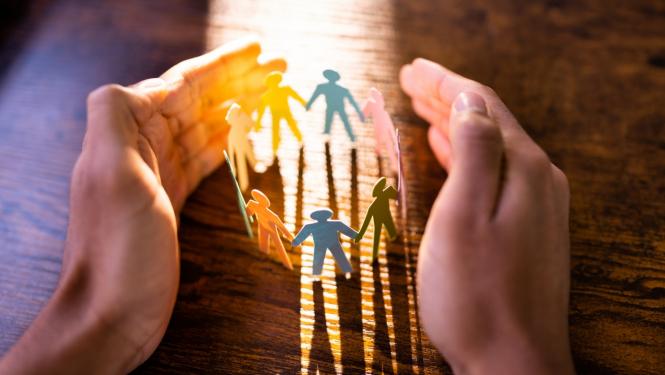 cilvēka plaukstas ir saliktas uz galda, veidojot aplīti iekšā starp plaukstām ir izgriezti no papīra cilvēciņi 