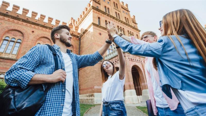 Jaunieši studenti sasit priecīgi plaukstas, fonā universitātes ēka