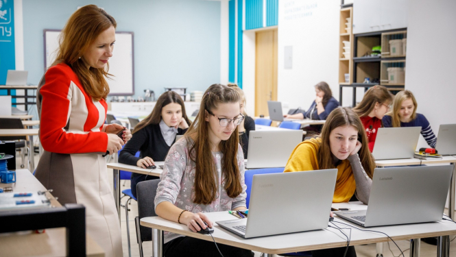 skolēni sēž klasē pie datoriem blakus stāv skolotāja