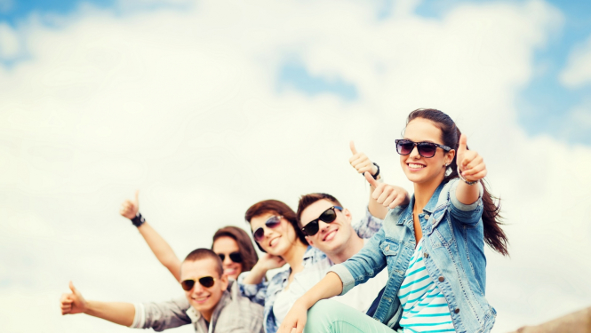 Jaunieši saulesbrillēs sēž uz zeme, smaida un rāda lielo īkšķi