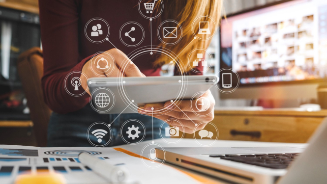 Darba galds ar klēpjdatoru, sieviete tur rokās planšeti, kurā attēlotas ikonas