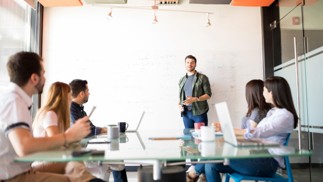 biroja telpā cilvēku grupa klausas vienu cilveku pie tāfeles