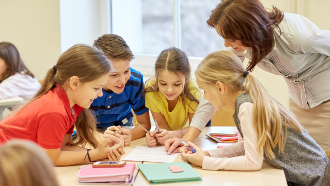 vairāki skolēni sēž grupā pie galda skolotāja stāv blakus un kaut ko norāda viņiem