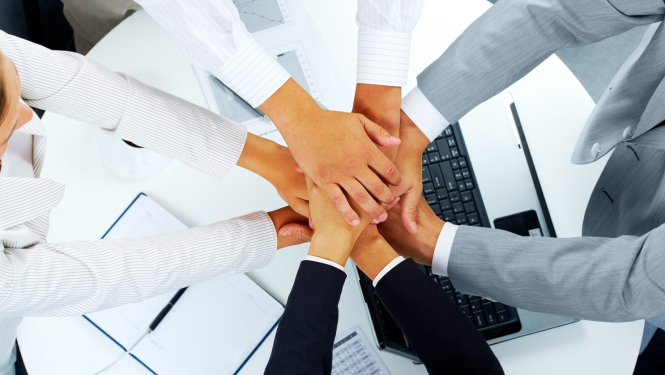 Četri cilvēki sadevušies rokās, fonā redzami dokumenti un klēpjdators