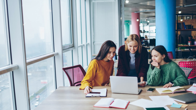 Trīs sievietes sēž pie galda un skatās datora ekrānā