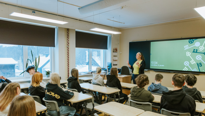 skolēni sēž pie soliem klasē, klases priekšā stāv skolotāja pie interaktīvās tāfeles 