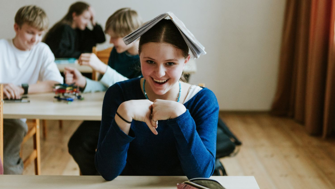 meitene skolniece sēž skolas solā un smaida, uz galvas viņai ulikta atvērta grāmata; aizmugurējā plānā sēž skolas solā citi skolēni