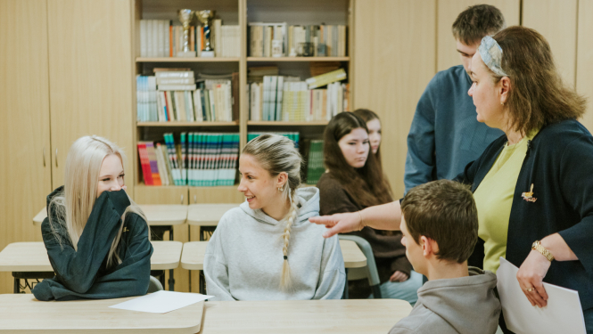 klasē solā sēž pusaudži un smejas, blakus stāv skolotāja un kaut ko runā