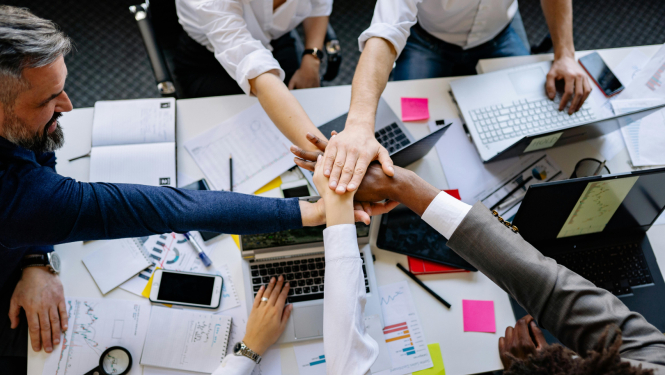 vairāki cilvēki sēž pie viena darba galda pa apli un pa vidu galdam sakruastoja rokas, uzliekot vienu otram virsū; uz galda ir dažādas pierakstu lapas, telefons, dators