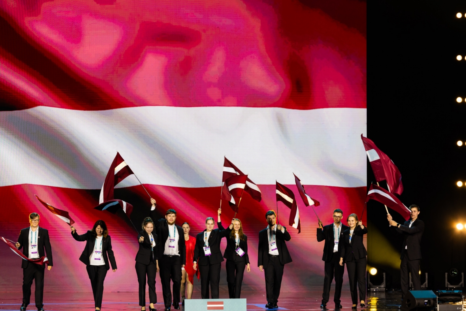 Latvijas komanda ar Latvijas karogiem un lielu karogu fonā