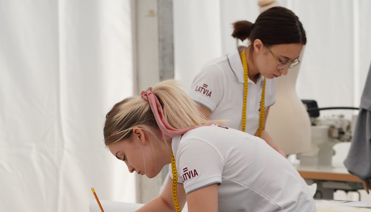 Meitene pie galda zīmē ar zīmuli, fonā redzama otra konkursante ar mērlenti ap kaklu